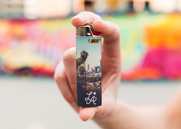 A lightskinned hand holding up a Bic lighter. The lighter is skinned with a photo of Los Angeles.