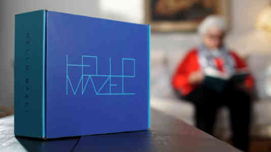 Out-of-focus photo of an old woman in red sitting on a couch. In the foreground is a Hello Mazel package.